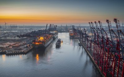Aerial view of a container terminal with large container cargo ships and gantry cranes. A cargo ship  is navigating through the central waterway.  On both sides of the waterway are massive container terminals lined with red and black gantry cranes used for loading and unloading ships.  on the left docked ships with brightly lit containers. Moody sky at dawn. Port of Hamburg in Germany, Europe.