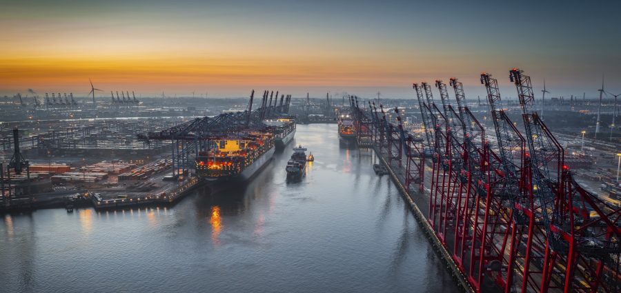 Aerial view of a container terminal with large container cargo ships and gantry cranes. A cargo ship  is navigating through the central waterway.  On both sides of the waterway are massive container terminals lined with red and black gantry cranes used for loading and unloading ships.  on the left docked ships with brightly lit containers. Moody sky at dawn. Port of Hamburg in Germany, Europe.