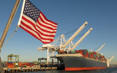 An American flag waves in the foreground at this US port, where a cargo ship loaded with containers is berthed beneath giant cranes on a clear blue sky day. The image symbolizes concepts like Made in America, Made in the USA, industry, global economy, pride, strength, power, patriotism, trade, manufacturing, and the idea of getting back to work.
