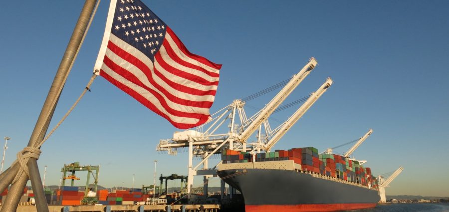 An American flag waves in the foreground at this US port, where a cargo ship loaded with containers is berthed beneath giant cranes on a clear blue sky day. The image symbolizes concepts like Made in America, Made in the USA, industry, global economy, pride, strength, power, patriotism, trade, manufacturing, and the idea of getting back to work.