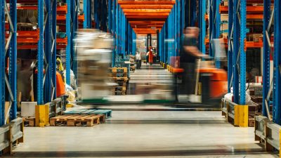 Warehouse workers are operating forklifts, transporting goods between tall shelves filled with various products, showcasing the fast paced environment of a modern distribution center