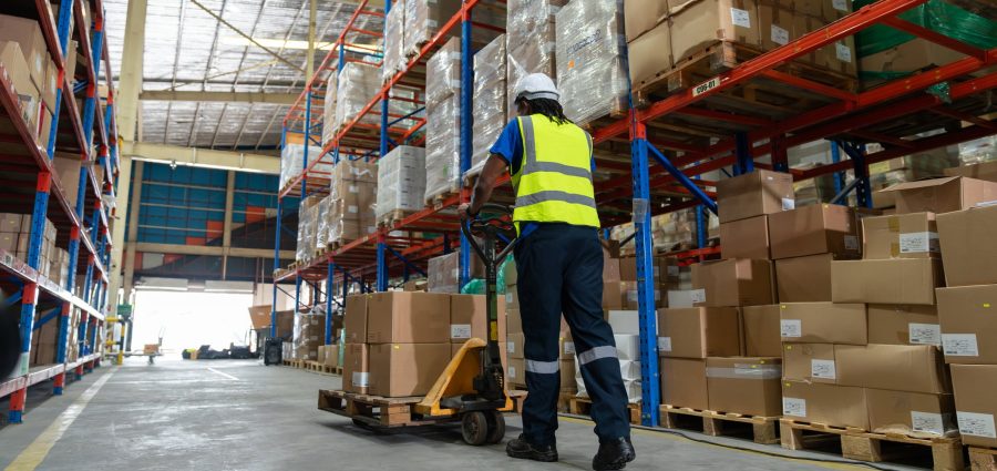 African American man worker using pallet truck for move cardboard boxes. Storehouse employee in uniform working and unloading goods in warehouse. Logistics, Distribution Center concept.