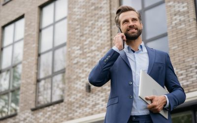 Handsome smiling businessman wearing stylish suit talking on mobile phone, holding laptop standing on the street. Successful business