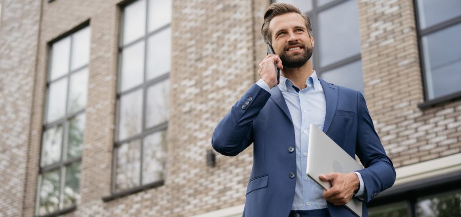 Handsome smiling businessman wearing stylish suit talking on mobile phone, holding laptop standing on the street. Successful business